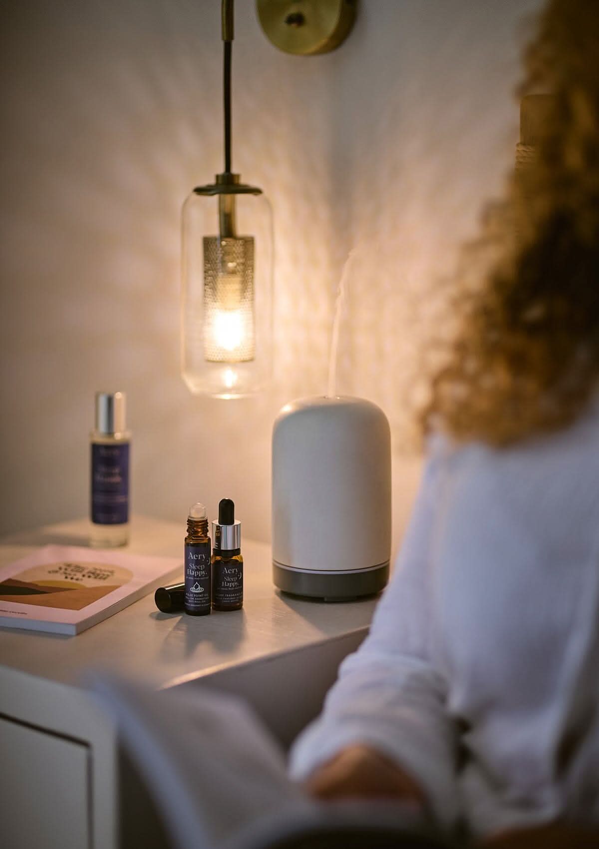 Blue Sleep Happy pulse point oil displayed next to Sleep Happy fragrance oil and pillow mist placed on bedside table with books and electric diffuser