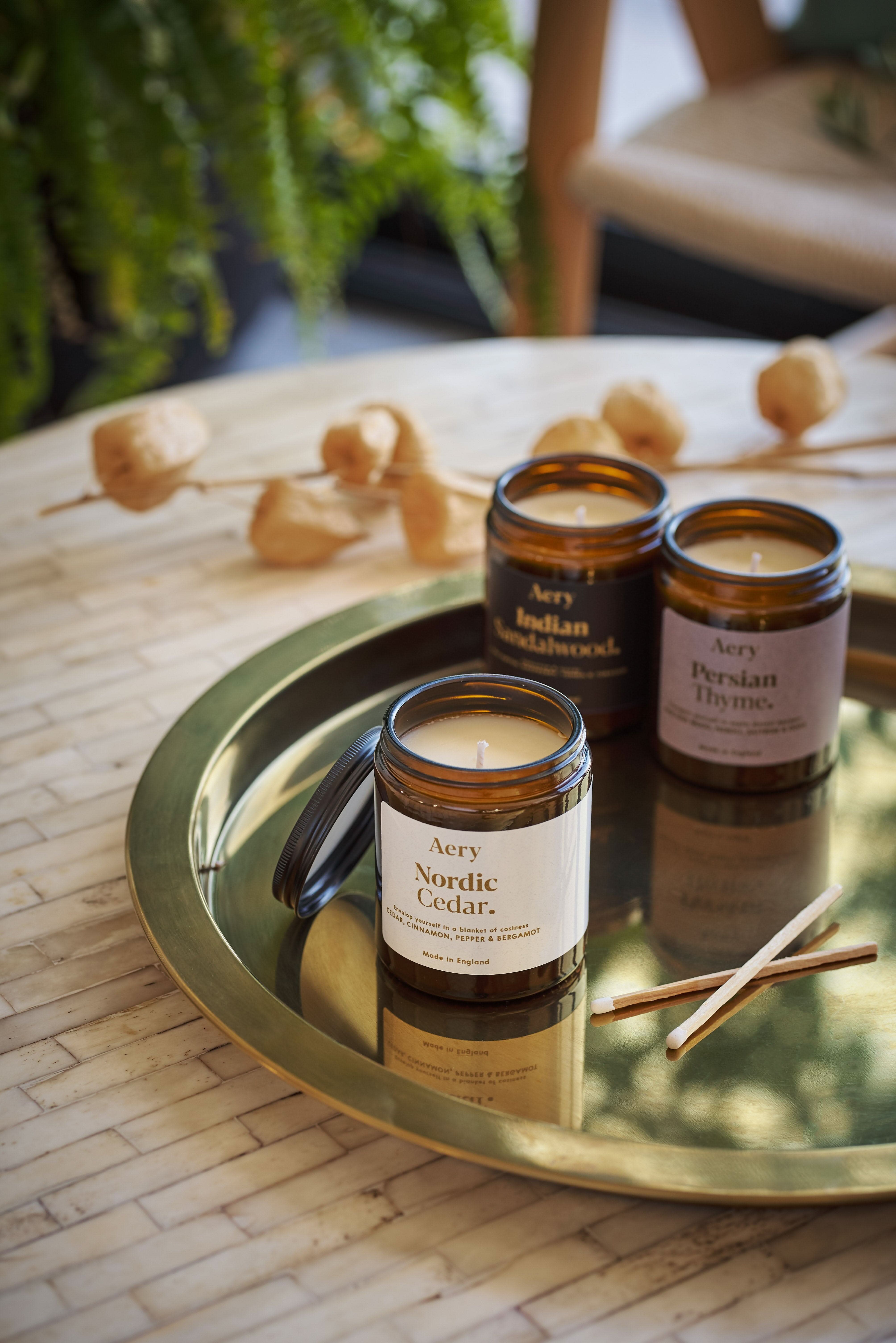White Nordic Cedar jar candle displayed next to Indian Sandalwood and Persian Thyme jar candles by Aery placed on a gold circle tray on cream table