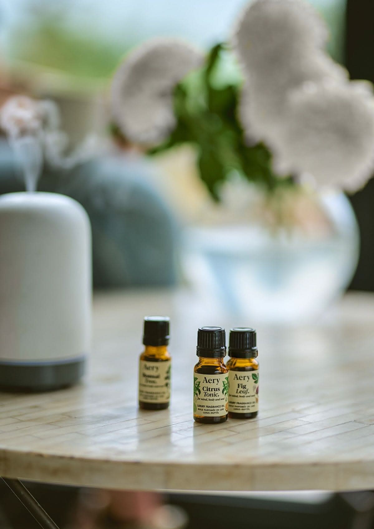 Cream Botanical set of three fragrance oils by Aery displayed next to electric diffuser and vase of flowers on round table