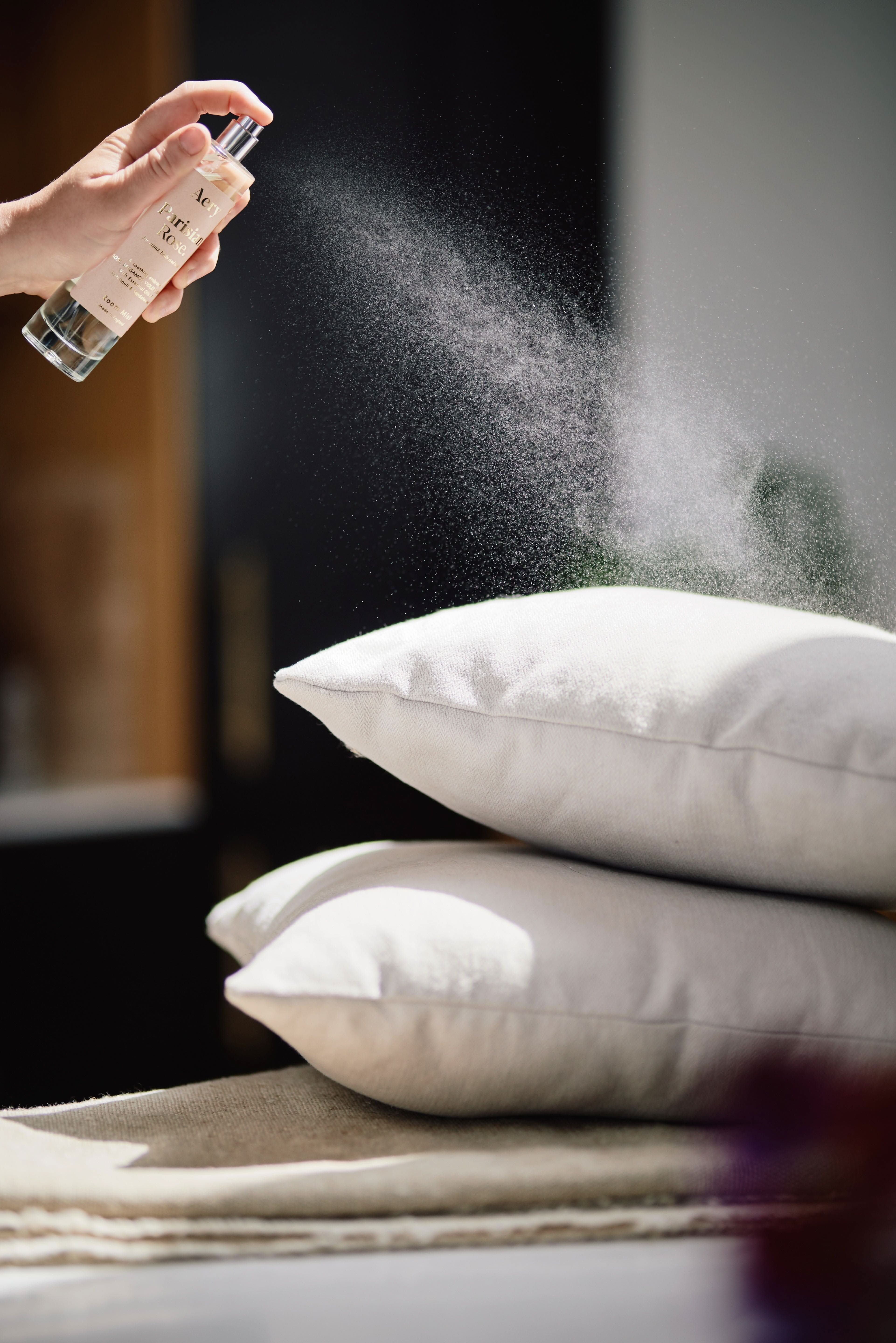 Pale Pink Parisian Rose room mist by Aery displayed in hand with grey cushions